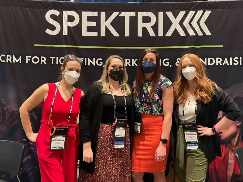 4 women wearing masks and conference lanyards in front of the Spektrix exhibit booth