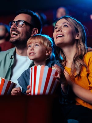 A family of three watch something at the theatre, holding red and white striped popcorn buckets.