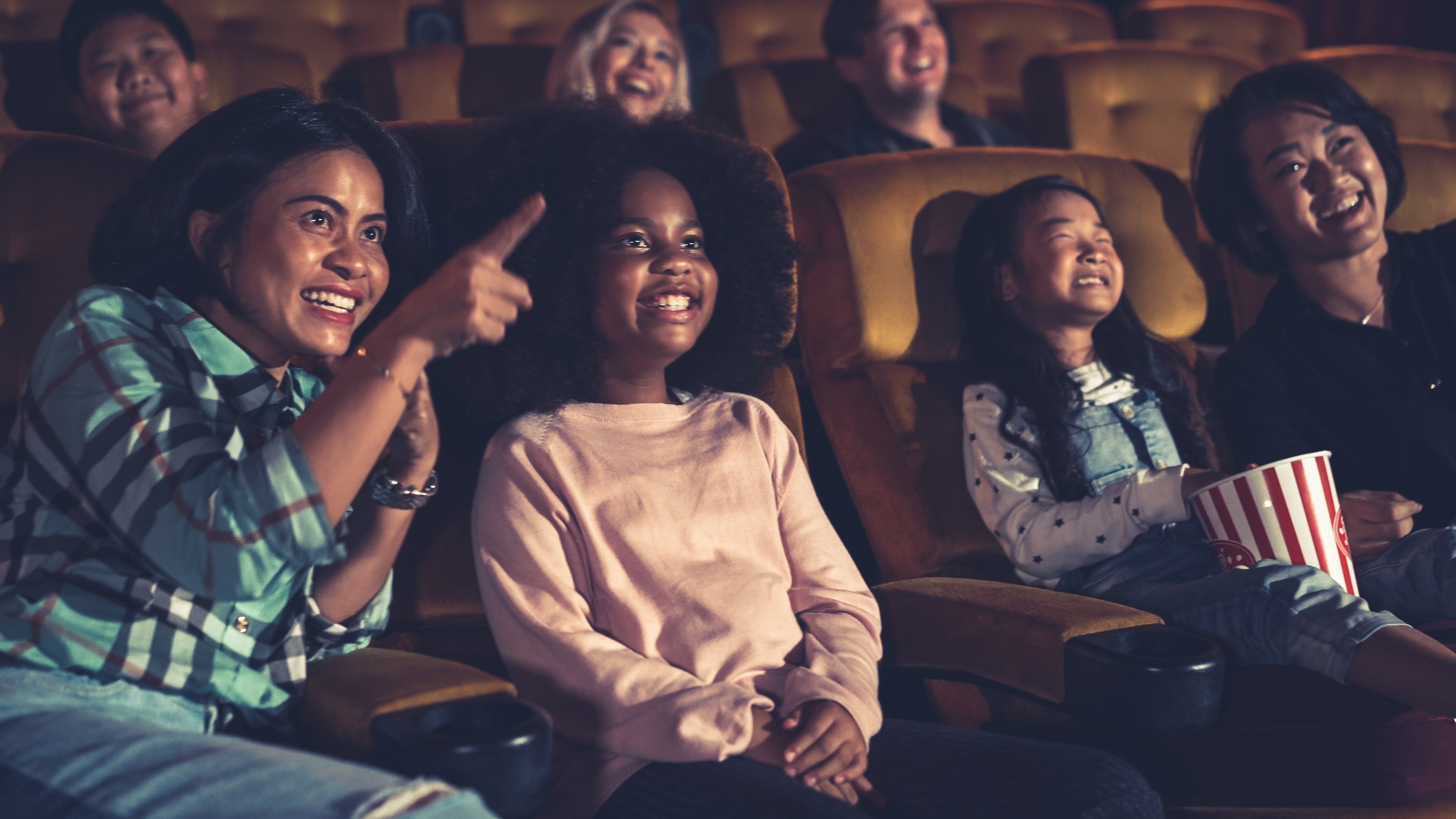 A multi-generational audience smiles and laughs while watching something at the cinema, reclining against yellow theatre seats.