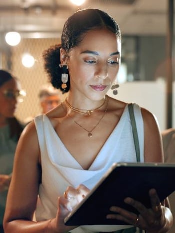 A woman in a white cowl neck tank top with drop earrings and a low ponytail uses a tablet.