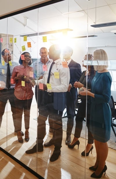 Coworkers gather around a board covered in sticky notes, smiling as the sun beams through behind them.