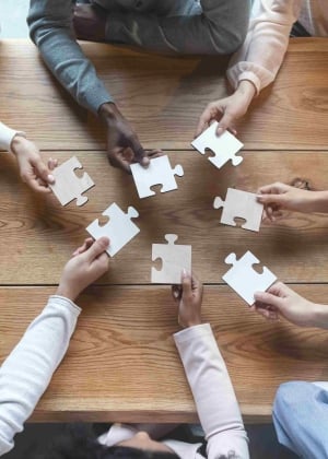 A group of people hold puzzle pieces above the center of a wooden table.