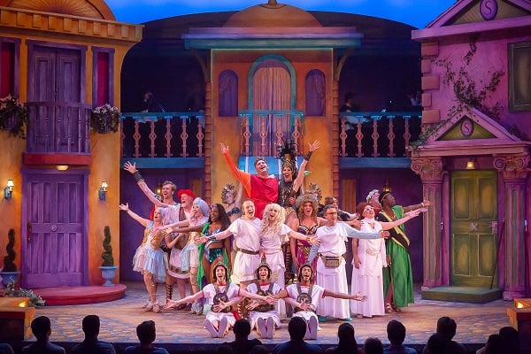 Performers in Roman-style toga costumes raise their arms in a finale, in front of colorful staging representing a street of houses