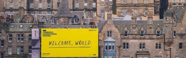 The grey-brown stone of Edinburgh rooftops, with a bright yellow sign saying 'Welcome, World'