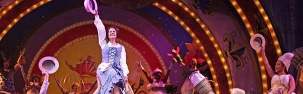 A girl in a blue pinafore dress is held aloft, waving a hat in triumph, by fellow performers at Music Theatre Wichita