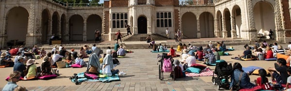 An audience gathered on picnic rugs in an arched stone courtyard on a sunny day, enjoying live outdoor opera