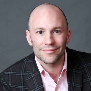A headshot of Eric Nelson, a man with a shaved head, wearing a light pink shirt and dark brown blazer that features blue and grey lines.
