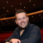 Josh Larson wears a black shirt and sits in a theater, smiling at the camera