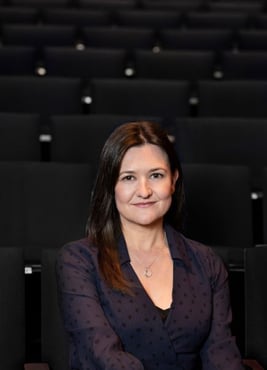 A headshot of Rani Haywood, a woman in her mid-forties with long dark hair wearing a navy dress and sitting in an empty theater