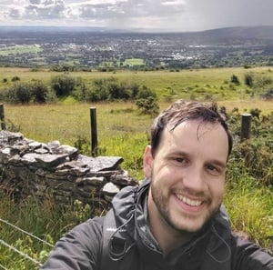 A selfie of Darko, a white male in his early 30's, with a nice country landscape background with partially cloudy skies taken in the Cotswolds. Darko has a toothy smile even though his short brown hair is wet from the rain. He is wearing a grey & black raincoat with black backpack straps as the picture cuts off just below his shoulders.