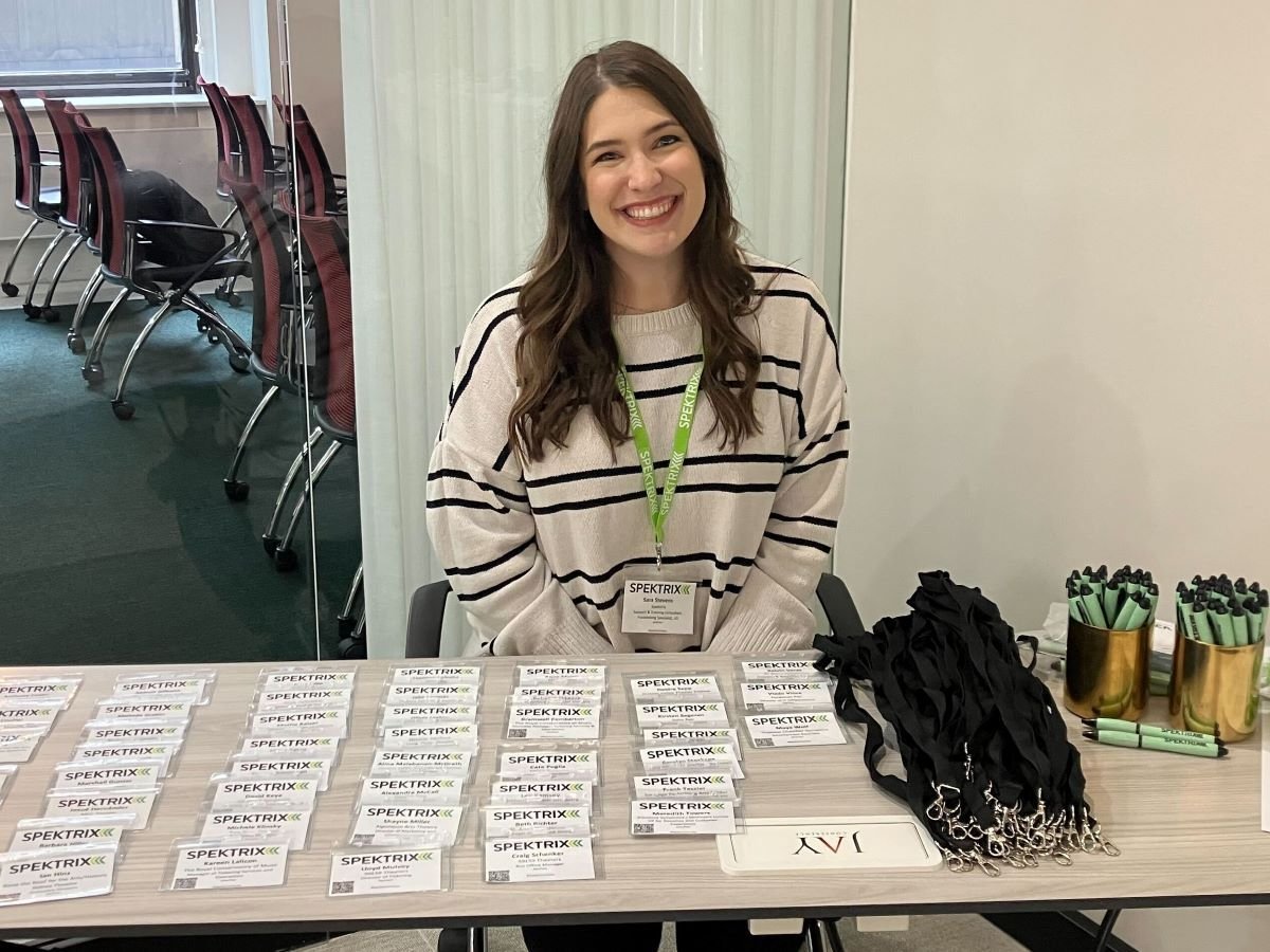 Sara Stevens, woman with long brown hair, sits smiling at a registration table with name tags and pens