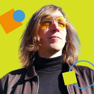 A mid shot of Matt Akers, a man with shoulder length light brown hear, wearing a black turtle neck and yellow tinted glasses, gazing into the distance