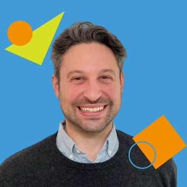 A headshot of Mike Geller, a white man with dark, salt and pepper, straight hair and a faint dark beard, wearing a black sweater over a blue collared shirt and smiling into the camera.