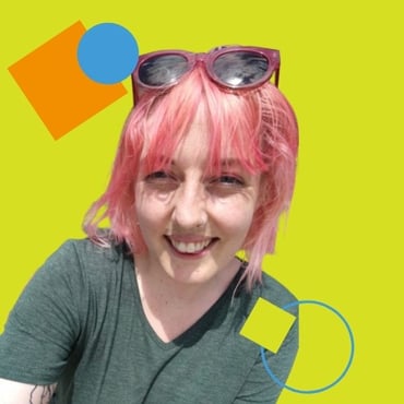 Lauren Watson wearing a green dress with pink hair and pink sunglasses is smiling at the camera. Their tattoo is also visible on their right arm.