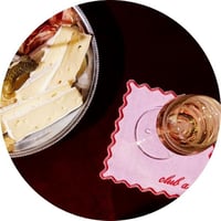 A plate of brie and pickles next to a glass of rose atop a pink cloth napkin with scalloped edges, a red border, and 