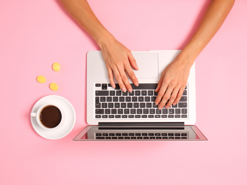 an image of hands typing at a laptop with a cup of coffee next to it on a pink surface