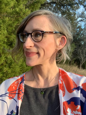 Headshot of Mindy Barenblat, a woman with short brown hair in glasses and colorful shirt standing outside, looking off camera, smiling.