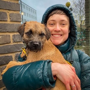 A headshot of Anne Harvey, a white woman with very short brown hair, wearing a hooded coat and holding a small dog.