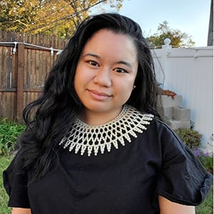 A headshot of Jessica  Abejar in an outdoor setting, wearing an elaborate pearl collar necklace.