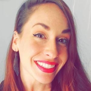 A headshot of Karen Elizondo, a woman with long straight brown hair, purple leaf-shaped earrings, and pink lipstick smiling into the camera.