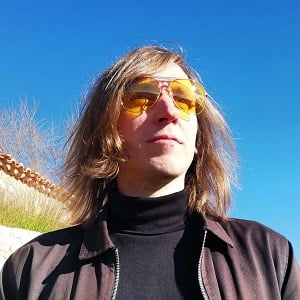 A mid shot of Matt Akers, a man with shoulder length light brown hear, wearing a black turtle neck and yellow tinted glasses, gazing into the distance with bright blue sky in the background.