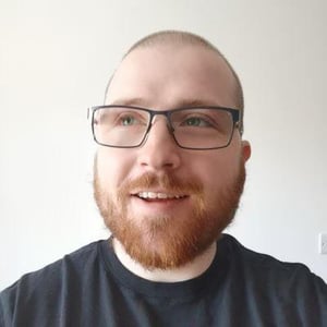A photo of AJ smiling. He has a very short shaved brown hair, glasses and a ginger beard.