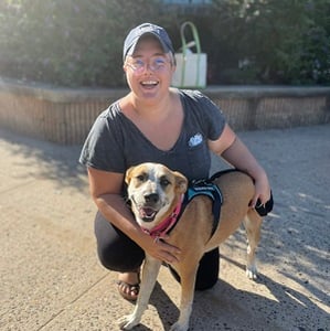 A sunny image of Sarah Caswell, a white woman wearing a hat and crouched next to her service dog, a medium sized fawn colored cattle mix with freckles, both have big smiles.