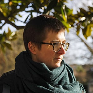 Aenne lotze, a person with short hair, glasses and wrapped in a green scarf stands below a tree, looking to the side