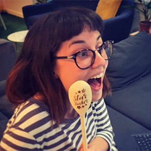 A white woman in her thirties with shoulder length brown hair and glasses, excitedly holding a Star Baker award spoon after winning Spektrix Bake-Off.