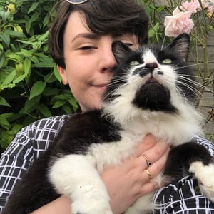 Annie Loake, a woman with short dark hair, hold a fluffy black and white cat in a garden while smiling at the camera