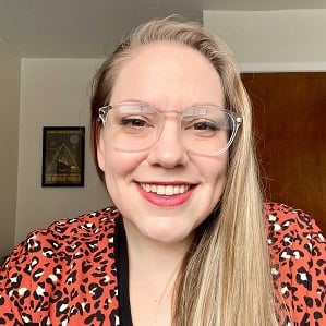 Bethany Nothstein, a white woman with blonde hair and glasses wearing a black shirt and leopard print jacket smiling at the camera.