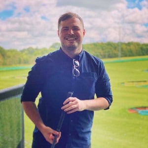 Billy Fluck, a man in his twenties, smiling and holding a golf club at a driving range.