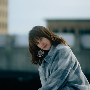 Charlie, a white person with shoulder-length brown hair and bangs, looks over their shoulder toward the camera wearing a grey and white wool coat.