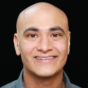 Headshot of James Menezes, a tan bald man in his early thirties smiling into the camera
