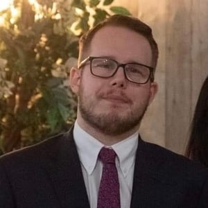 James has short, dark hair and short facial hair and is wearing a suit and glasses.