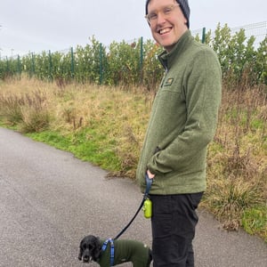 James stands in field, trying to keep his small dog under control. He wears a green jacket, glasses, and a hat.