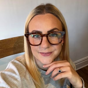 A headshot of Jennifer Caveney,  a woman with blonde straight hair wearing a grey jumper and glasses and smiling into the camera.