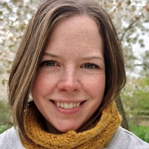 A headshot of Keren Nicol, a woman in her 30s with brown hair and freckles wearing a grey sweater and mustard-coloured knitted snood. She's outside with blossom trees around her.