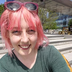 Lauren Watson wearing a green dress with pink hair and pink sunglasses is smiling at the camera. Their tattoo is also visible on their right arm.