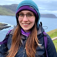 Lexi Messenger, a woman in her 30's, in hiking wear, standing on the coast of the Isle of Skye and smiling to the camera.