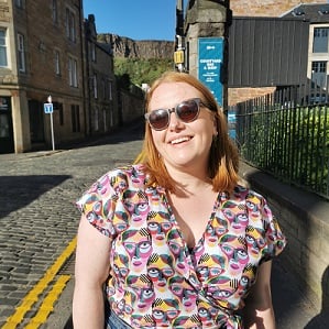 Miriam Wright, a woman with bobbed red hair, outside in the sunshine. Smiling with sunglasses and a patterned shirt.