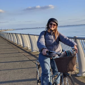 Rachael is pictured riding on a blue bicycle wearing a helmet. The bicylce has a basket with a blanket and Rachael is riding along the waterfront in Liverpool.