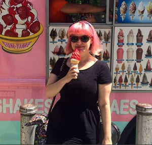 Victoria, a woman with pink hair is standing in front of an ice cream van, wearing a black dress, sunglasses and eating a giant pink ice cream.
