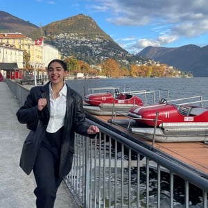 Emily walks along a lake smiling, dark hair pulled back in a bun, wearing a black coat, white collared shirt, and black slacks.