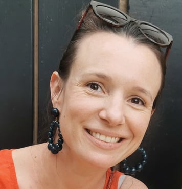 A headshot of Raphaelle Roux, a white woman with brown hair, wearing an orange top and large earrings and smiling into the camera.