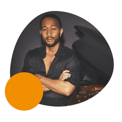 john legend standing with his arms crossed next to a piano against a dark wall