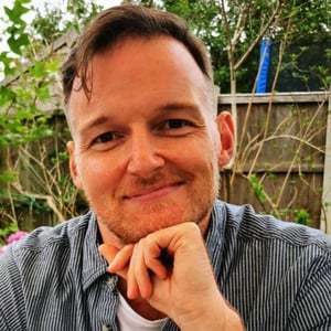 Allan Foy sat at a table with his hard resting under his chin wearing a striped shirt and smiling.