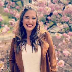 A photo of Sara, a white woman in her late twenties with brown wavy hair, in front of the cherry blossoms at the Brooklyn Botanical Gardens.