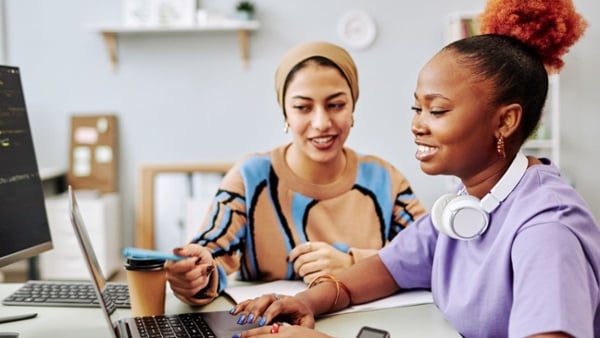 two young women working on project with a laptop together in an office and smiling