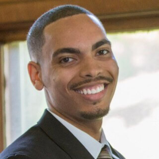 Zack Moore, a Black man, wearing a suit and tie and smiling into the camera. (1)